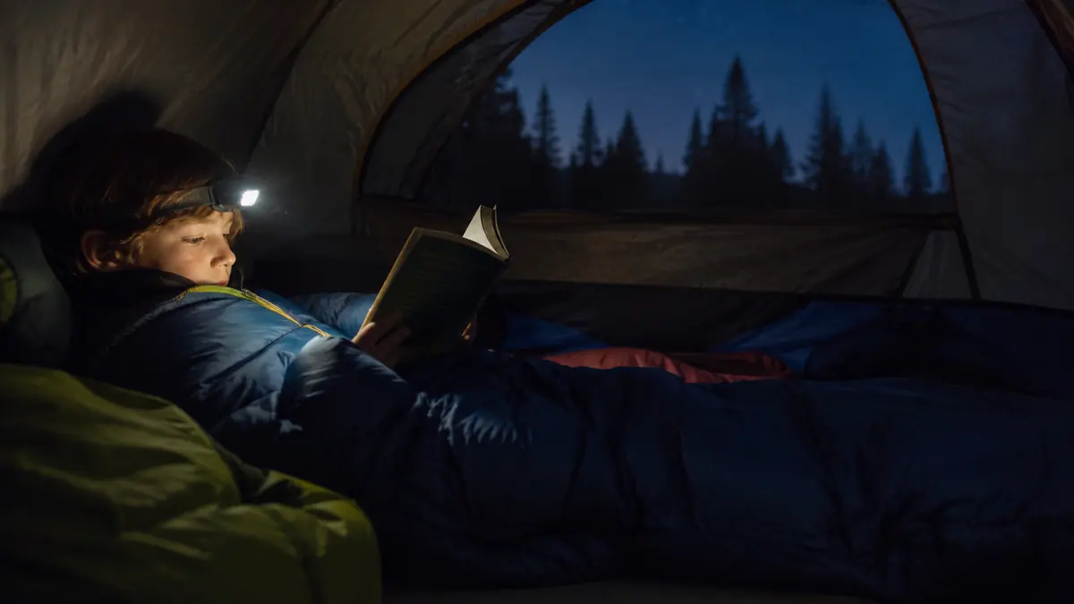 A kid zipped into a sleeping bag reading with a headlamp inside a tent