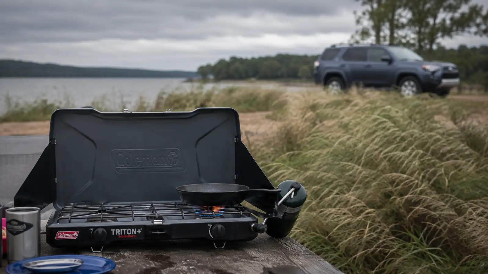 Coleman Triton+ camp stove burning steadily on a windy morning at a lakeside campsite
