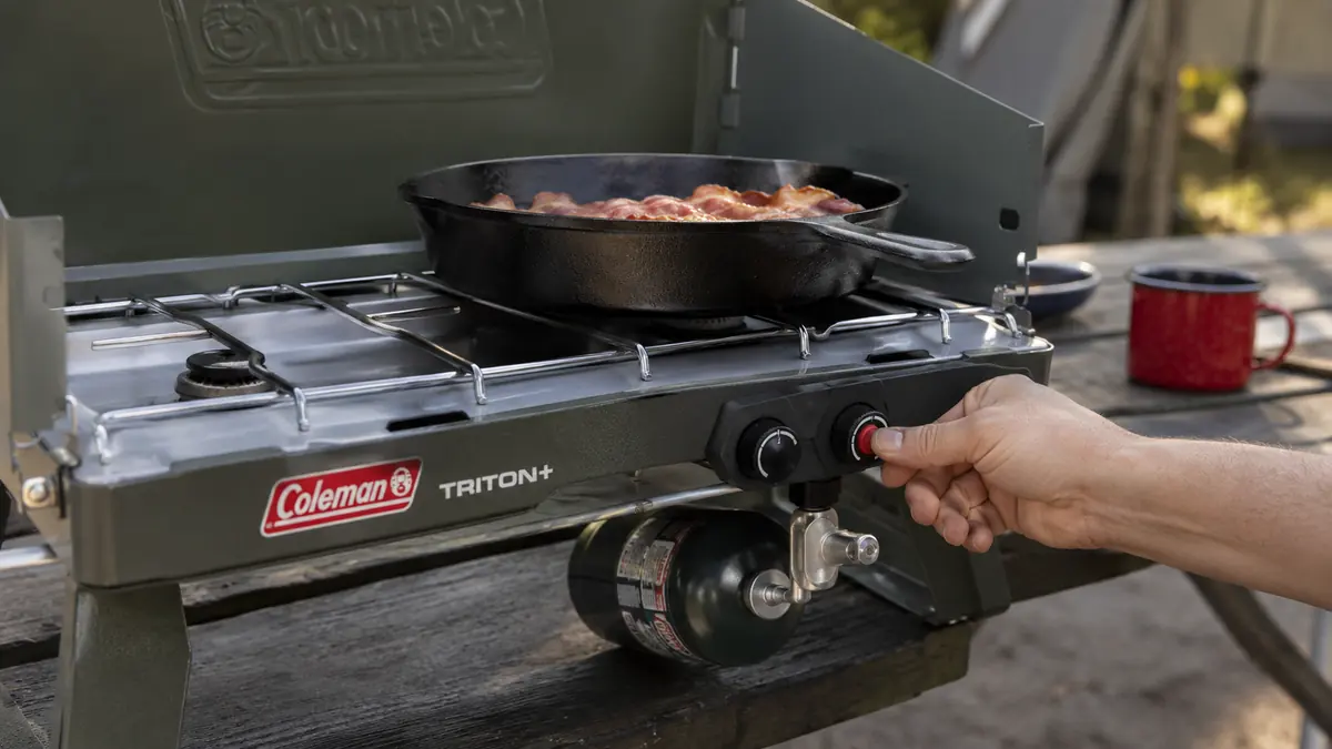 Hand pressing the InstaStart ignition button on the Coleman Triton+ stove with a skillet of bacon on the other burner