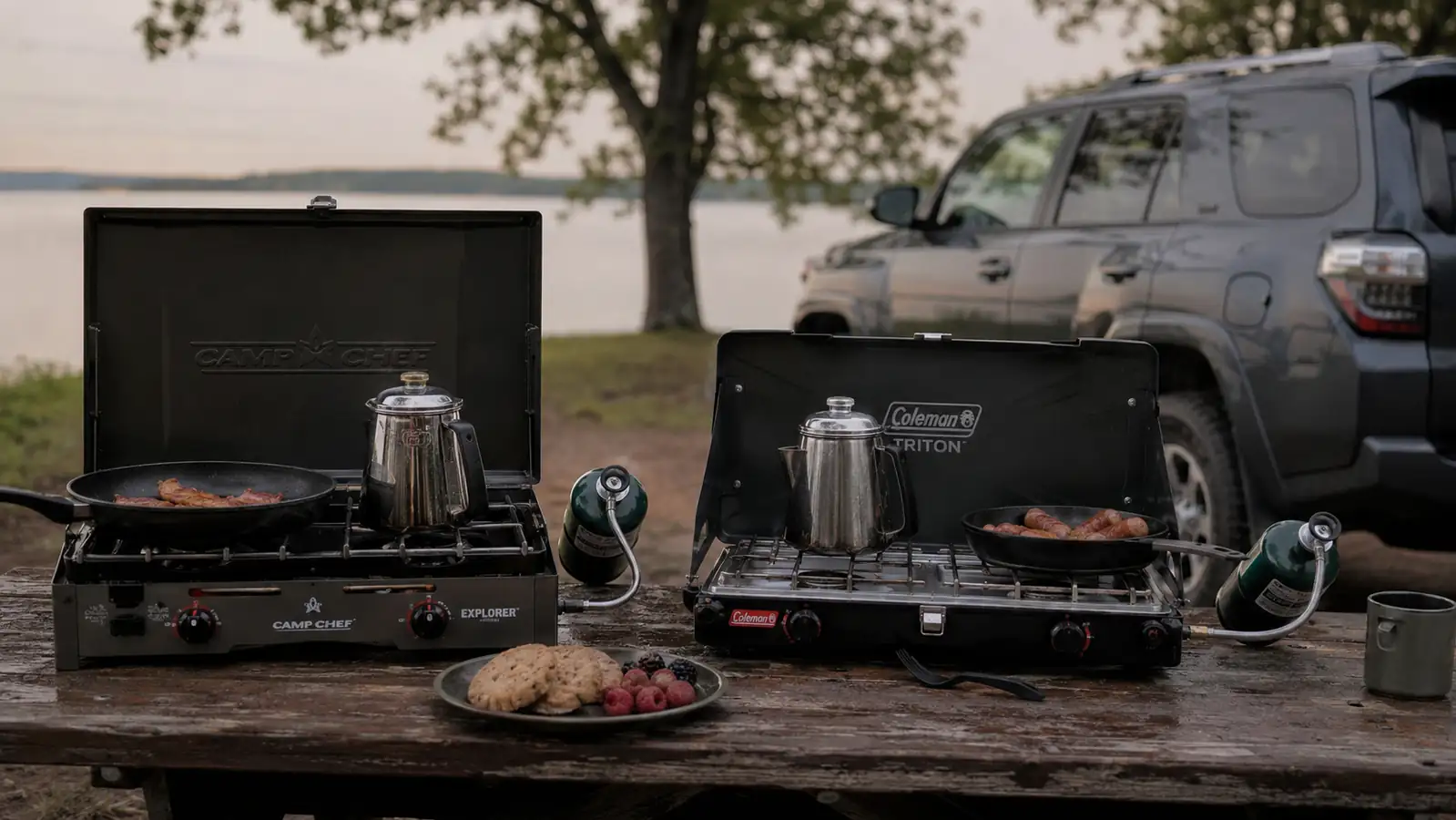 Coleman Triton+ 2-burner stove and a Camp Chef Explorer stove set up side by side on a picnic table at a lakeside campsite