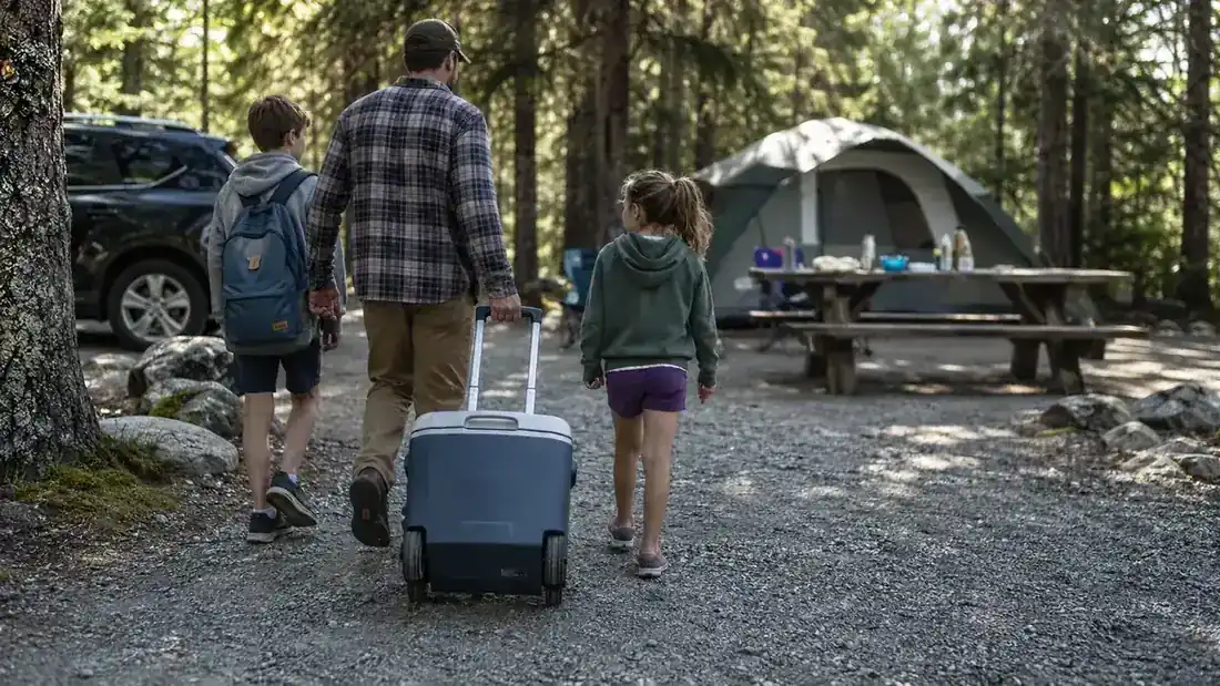 A rolling cooler being pulled by its handle up a gravel path toward a campsite with a tent visible ahead