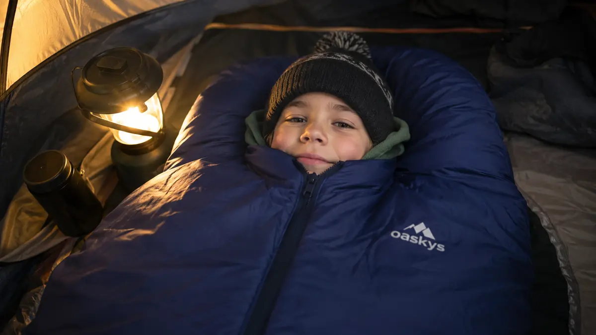 Kid zipped into the oaskys sleeping bag wearing a fleece layer, only face visible, tucked in for a cold night