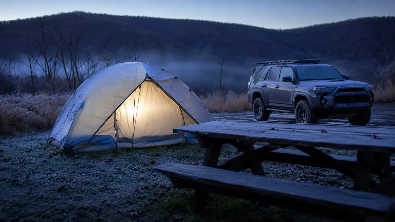 Frosty morning campsite with a tent glowing softly from a headlamp inside, breath visible in the cold air