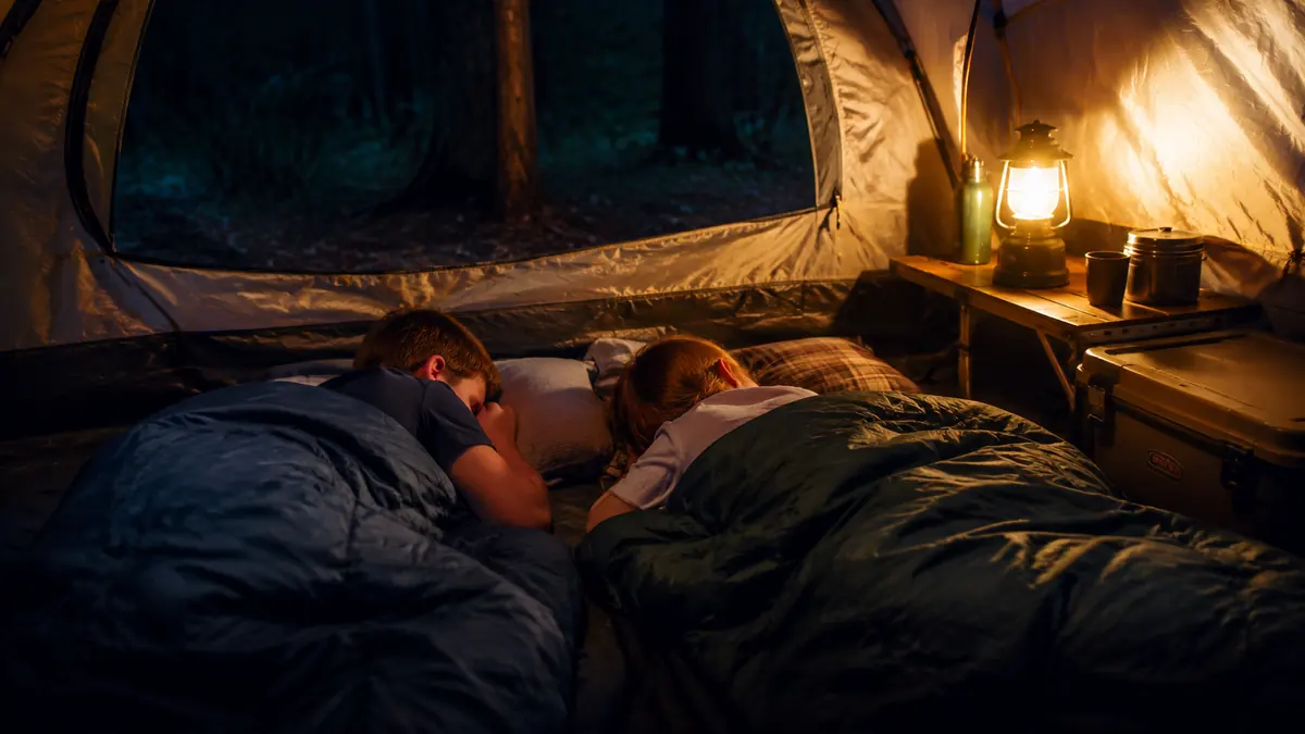 Two kids asleep in sleeping bags inside a tent lit by a warm lantern glow