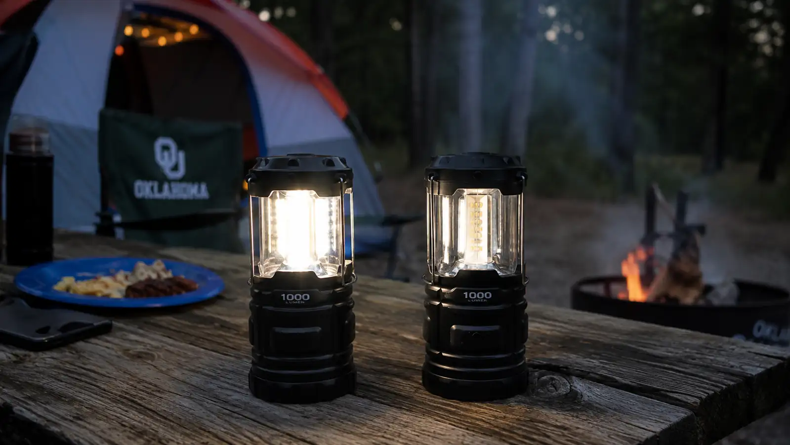 Two rechargeable camping lanterns sitting side by side on a weathered picnic table at dusk, one glowing noticeably brighter than the other