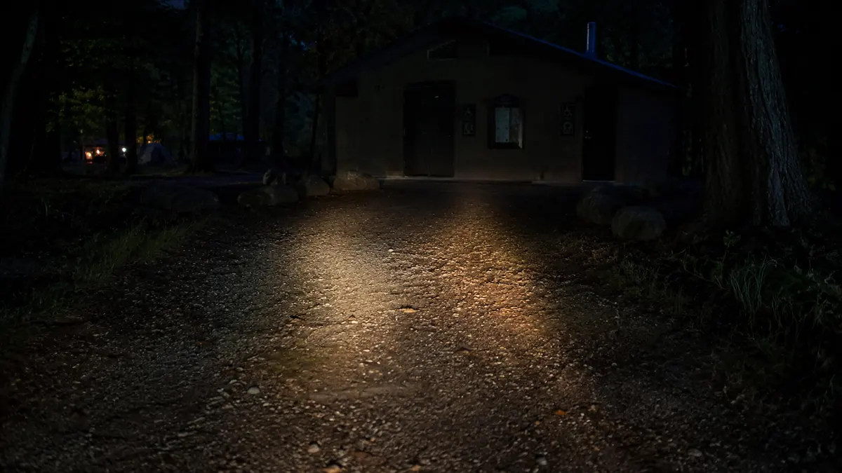 A lit bathhouse path at a campground with a person walking a flashlight beam ahead of them at night