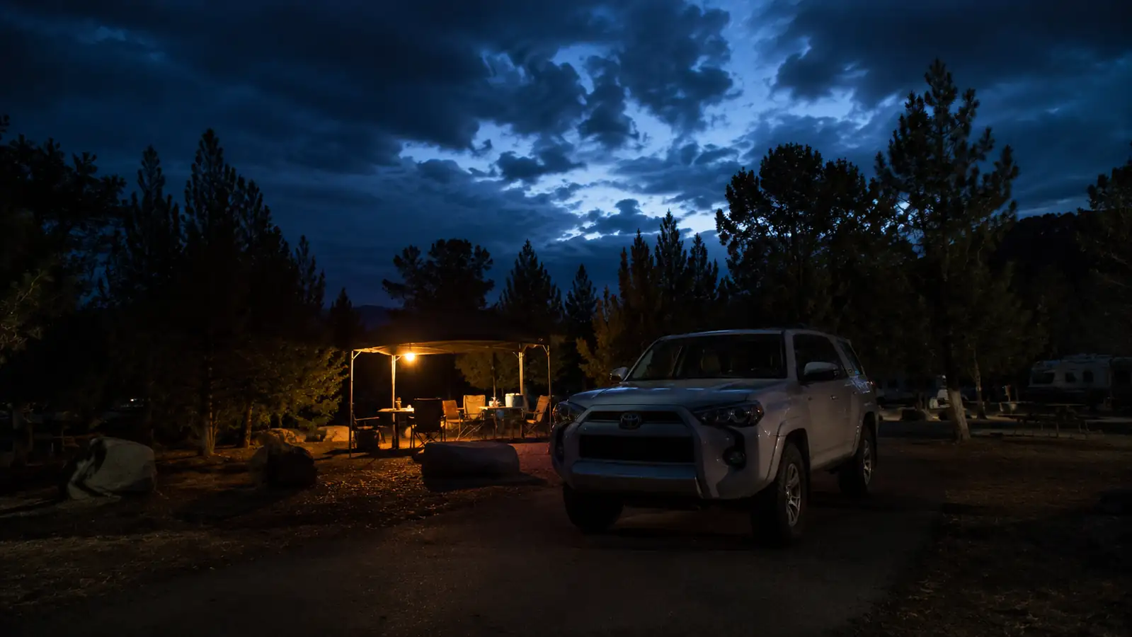 A dark campground loop with one lit campsite glowing warmly from a lantern while the rest of the loop sits in shadow